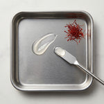 Metal tray with saffron threads, a droplet of liquid, and a metal spoon on a light gray background.