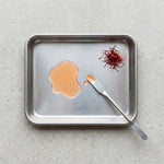 Metal tray with saffron threads and a dollop of liquid on a light gray background