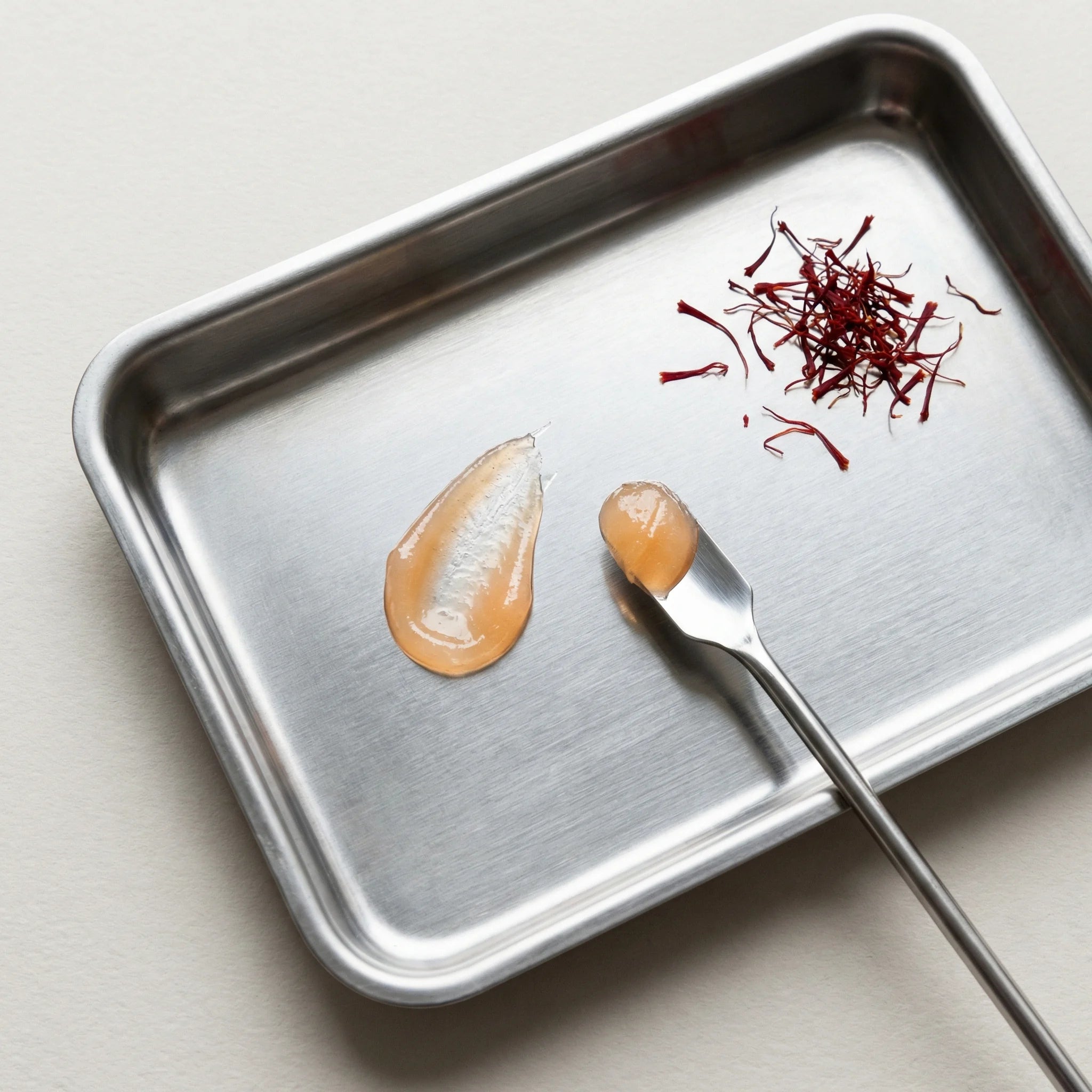 Metal tray with saffron threads and two droplets of lip oil on a light background