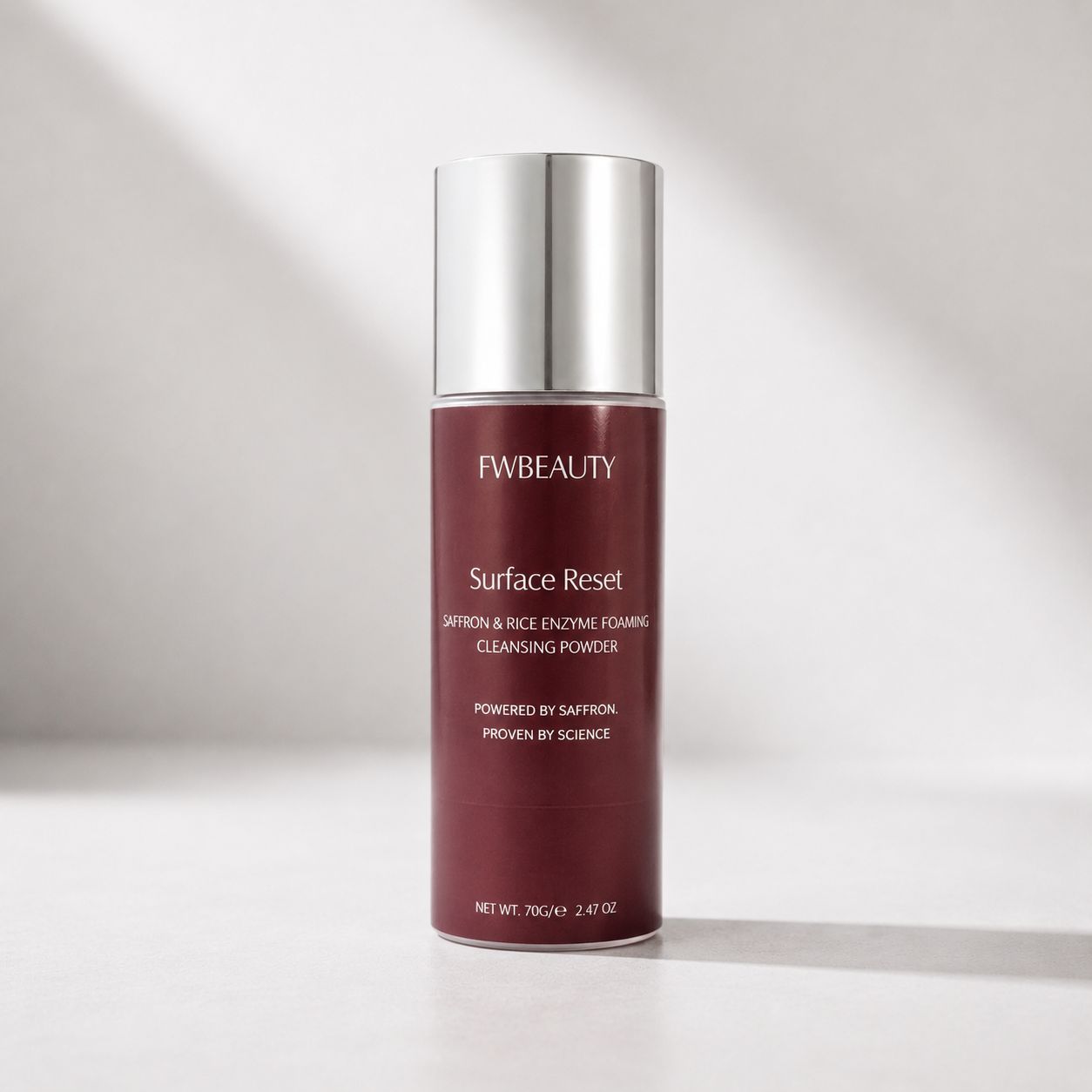 FWBEAUTY Surface Reset Cleansing Powder in a matte burgundy bottle with a reflective silver cap, set against a warm, softly lit gray background.